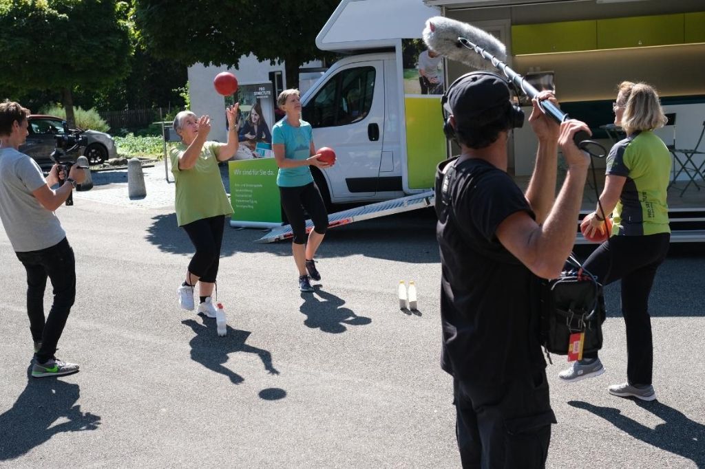 Vor dem Infobus wird ein Aktivierungsspiel gespielt und von einem Kamerateam festgehalten.