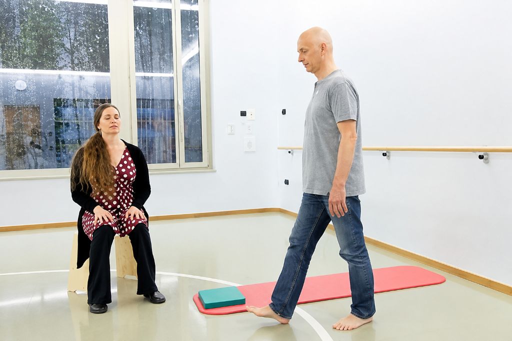 Senior führt in einer Turnhalle unter Aufsicht einer Mitarbeiterin sportliche Übungen aus.