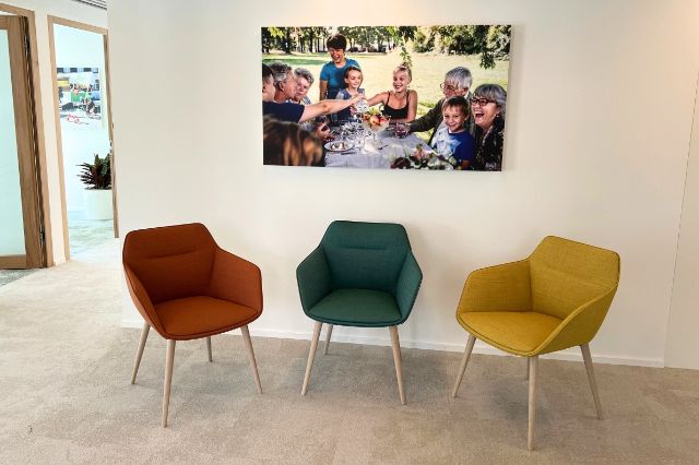 Auf dem Bild sehen wir drei bunte Stühle und ein grosses Bild an der Wand hängen.
