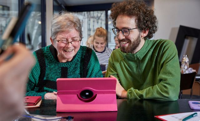 Ein junger Mann erklärt einer Seniorin die Funktionen auf ihrem Tablet.
